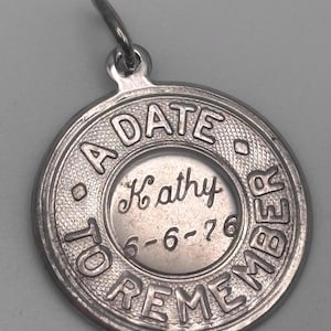 May include: A silver-colored circular pendant with the words "A DATE TO REMEMBER" around the edge. The name "Kathy" and the date "6-6-76" are in the center. The pendant has a small loop at the top for a chain.