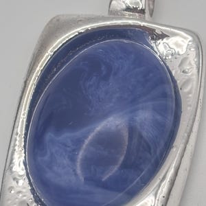 May include: A silver-toned pendant with a rectangular frame and a blue oval center. The blue center has a marbled appearance. The pendant has a textured border and a loop at the top for a chain.