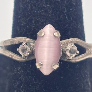 May include: A silver ring featuring a light pink, oval-shaped center stone with a cat's eye effect. Two smaller, clear stones flank the center stone. The ring has a split band design.