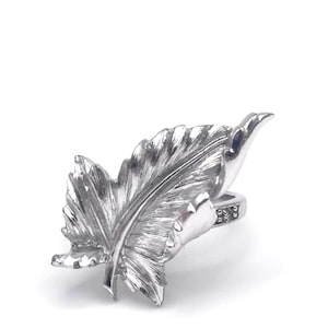 May include: A silver-colored ring featuring a detailed leaf design. The ring has a textured surface, with the leaf's veins and edges clearly defined. The ring's band has a decorative pattern.