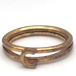 May include: A gold-toned metal ring featuring a double-band design that converges at the front. The front section displays a prominent letter C, which connects the two parallel bands. The metal surface shows a slightly weathered, matte finish.