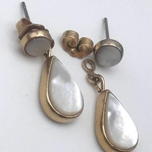 May include: A pair of gold-toned earrings with pear-shaped, white mother-of-pearl drops. The earrings have a post and a round, white stone at the top. A gold earring back is also present.