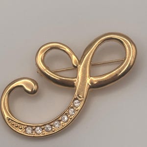 May include: Gold-toned brooch in a cursive "J" shape, adorned with sparkling clear crystals along the curve. The brooch has a pin closure on the back and a polished finish, suitable for adding a touch of elegance to any outfit.