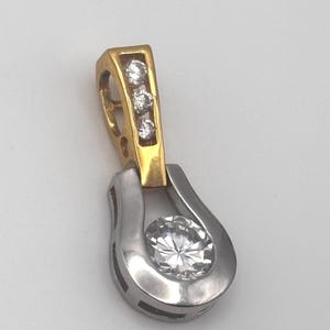 May include: A two-tone pendant with a gold-colored top section and a silver-colored bottom section. The gold section features three small, clear gemstones. The silver section holds a larger, round, clear gemstone. The pendant is designed to be worn as jewelry.