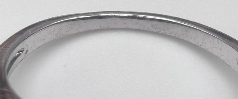 May include: Close-up of a silver ring band. The ring has a smooth, slightly curved surface with minor wear and tear visible. The band is a light, metallic color, reflecting the surrounding light.
