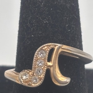 May include: A gold-toned ring featuring the letter "A" embellished with clear, round gemstones. The ring is set against a dark background, highlighting its design. The ring's band curves gracefully, adding to its elegant appearance.