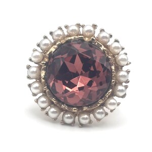 May include: A gold ring with a large, faceted, dark purple gemstone surrounded by a ring of white faux pearls.