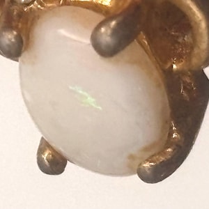May include: Close-up of a vintage gold-toned jewelry piece featuring a large, oval, iridescent white stone. The stone is held in place by four gold-toned prongs. The piece has a classic, elegant design.