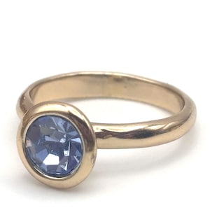 May include: A gold-toned metal ring featuring a smooth, polished band and a single, round, faceted periwinkle blue gemstone set in a circular bezel setting. The stone is positioned at the front of the ring, showcasing a brilliant cut.