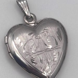 May include: Silver heart-shaped locket pendant with a detailed engraving of two birds perched on a branch. The locket has a textured surface and a bail for a chain. A romantic and sentimental piece of jewelry.