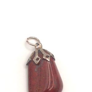 May include: A red-brown gemstone pendant with a silver-colored metal cap and loop. The pendant is a smooth, elongated shape with a polished surface. The metal cap has a decorative design.