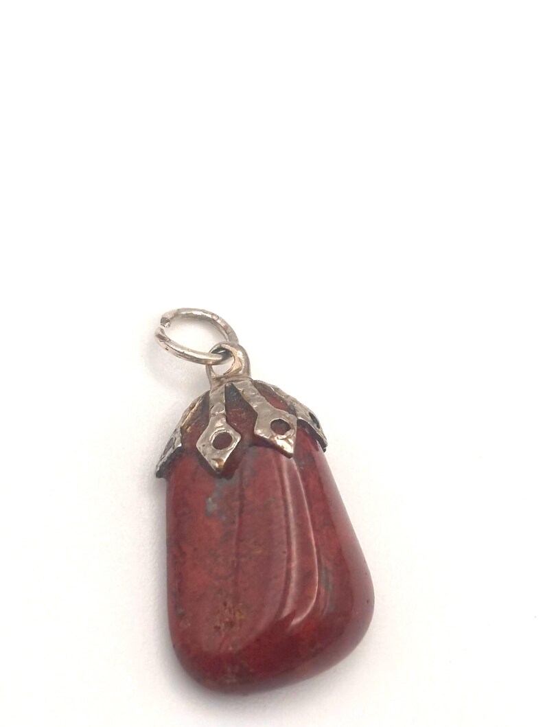 May include: A red-brown gemstone pendant with a silver-colored metal cap and loop. The pendant is a smooth, elongated shape with a polished surface. The metal cap has a decorative design.