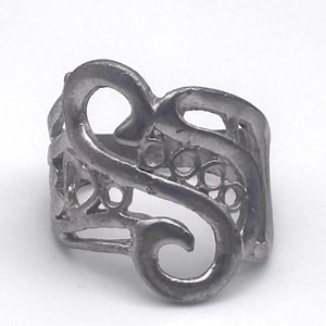 May include: A silver-colored ring with an openwork design. The ring features a swirling, abstract pattern with rounded shapes and cut-out details. The ring is made of a solid metal and has a textured appearance.