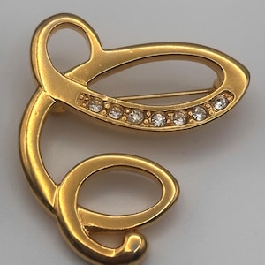 May include: Gold-toned brooch with a swirling, abstract design. The brooch features a series of clear, sparkling rhinestones set in a curved line. The pin is a decorative accessory.