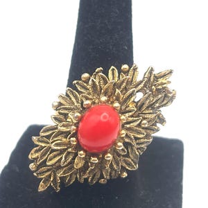 May include: A gold-toned ring with a red oval-shaped stone at the center. The ring features a detailed floral design with leaf-like elements surrounding the stone. The ring is displayed on a black velvet surface.