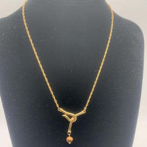 Pu&ograve; includere: Una collana con catena dorata con un ciondolo annodato e una piccola perla marrone. La catena &egrave; di stile corda.