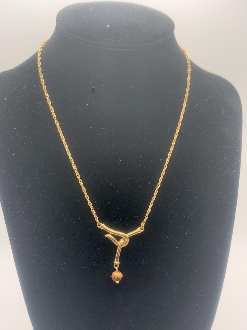 Pu&ograve; includere: Una collana con catena dorata con un ciondolo annodato e una piccola perla marrone. La catena &egrave; di stile corda.