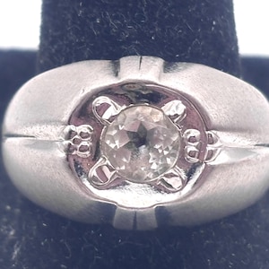 May include: A silver ring with a single, clear, round gemstone set in a raised, ornate design.