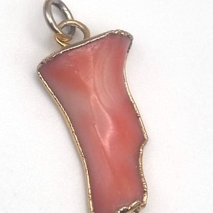 May include: A pendant featuring a smooth, irregular piece of salmon-colored coral with a polished finish. The coral is encased in a thin, textured gold-toned metal border and is attached to a small, circular silver-toned metal jump ring bail.