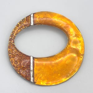 May include: A circular brooch featuring a vibrant orange enamel finish. One side displays a textured, hammered metal surface with a deep copper tone, while the other side is smooth and glossy. The two sections are separated by thin silver-toned metal bars.