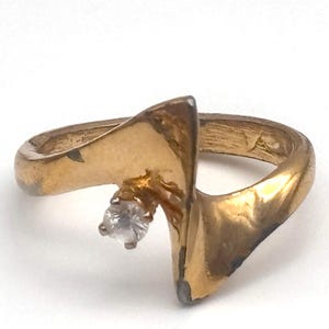 May include: A gold-toned ring featuring a fluid, asymmetrical band design that curves upward to a point. A single, small, clear, round-cut gemstone is set at the base of the central peak, contrasting with the metallic finish of the band.