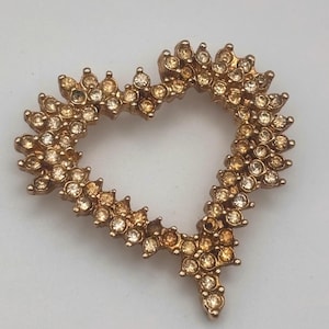 May include: A gold-toned heart-shaped brooch, encrusted with small, round, amber-colored rhinestones. The brooch has a textured, open-center design, with a pointed bottom. The rhinestones are arranged closely together, creating a sparkling effect.