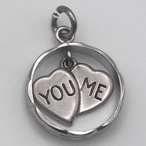 May include: A silver charm pendant featuring two heart-shaped charms within a circular frame. The hearts are inscribed with the words "YOU" and "ME". The pendant has a loop for attachment.