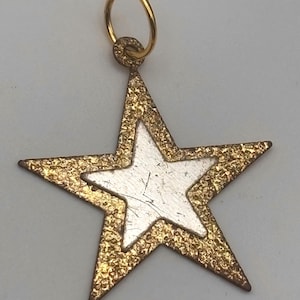 May include: A gold-colored star pendant with a textured border and a smaller silver-colored star in the center. The pendant has a small gold ring at the top for hanging.