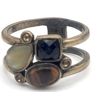 May include: A double-banded ring in a muted metallic finish featuring a cluster of stones. The arrangement includes a square black faceted stone, an oval brown tiger's eye, a pear-shaped iridescent white stone, and two small clear round accent stones.