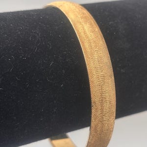 May include: A gold-colored bangle bracelet with a textured surface. The bracelet is displayed on a black velvet background, showcasing its metallic sheen. The bangle's design is simple and elegant, suitable for everyday wear.