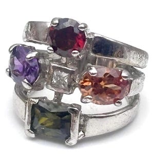 May include: A cluster of silver rings, each adorned with a different gemstone. The gemstones include a deep red oval, a purple oval, a square white stone, a peach-colored oval, and a green rectangular stone. The rings are stacked together.