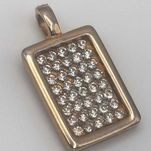 May include: A rectangular pendant with a gold-colored frame and bail. The center of the pendant is filled with numerous clear, sparkling crystals. The pendant is designed to be worn as a necklace.