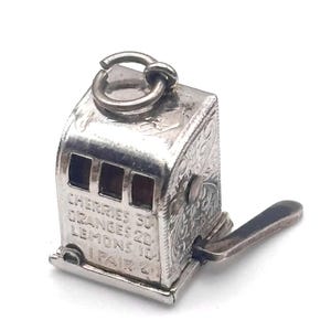 May include: A silver-toned miniature slot machine charm with etched floral designs. The charm has a pull lever and a ring for attachment. The text "CHERRIES 30, ORANGES 20, LEMONS 10, 1 PAIR 2" is visible on the front.