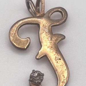 May include: A gold-toned pendant in the shape of the letter "f" with a small, round, clear gemstone at the bottom. The pendant has a loop at the top for a chain. The metal has a slightly aged appearance.