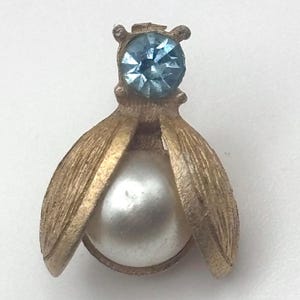 May include: Vintage gold-tone metal bee brooch with a light blue rhinestone head and a large, round, white pearl body. The wings are open, revealing the pearl. A classic, elegant jewelry piece.