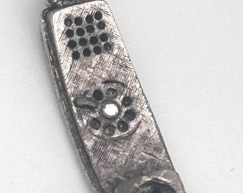 May include: A silver-toned metal charm shaped like a vintage telephone handset. The surface features a textured, brushed finish with a raised rotary dial, a speaker grid at the top, and a mouthpiece at the bottom. A small jump ring is attached at the top.