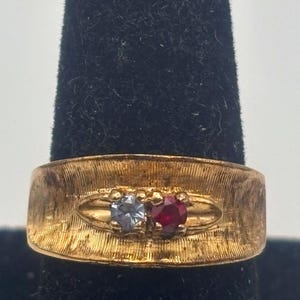 May include: Gold-toned ring with a textured band, featuring two gemstones: a light blue stone and a red stone. The ring is set against a dark background, highlighting its design and color.