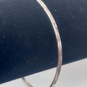 May include: A silver bangle bracelet with a textured, diamond-patterned design. The bracelet is thin and circular, with a polished finish. The bangle is displayed against a dark background.