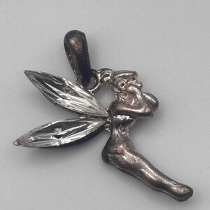 May include: A silver-toned fairy pendant with clear, faceted wings. The fairy is depicted in a seated pose, with detailed facial features and flowing lines. The pendant has a loop for a chain.