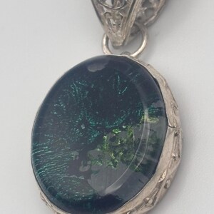 May include: A silver pendant with an oval-shaped green and black glass stone. The pendant has a filigree design around the stone and a bail at the top.