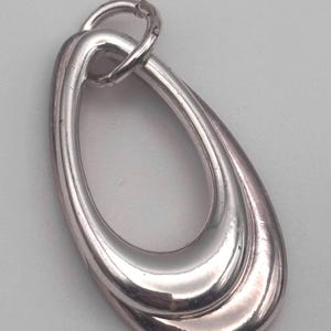 May include: A polished silver-toned pendant featuring two interlocking, smooth, teardrop-shaped loops. The pendant has a reflective metallic surface and includes a small circular jump ring attached at the top for hanging on a chain or cord.