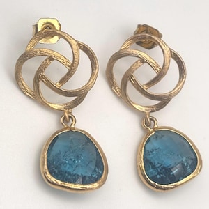 May include: A pair of gold-colored earrings. Each earring features a textured, intertwined knot design at the top, with a teardrop-shaped, blue gemstone pendant. The gemstones are set in a gold bezel.