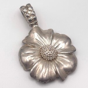 May include: A silver-toned flower pendant with a detailed petal design and a textured center. The pendant has a loop at the top for a chain. The flower pendant is a jewelry item.