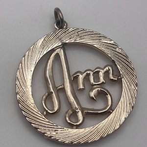 May include: A gold-toned pendant with the name "Ann" in a cursive font. The letters are within a textured, circular frame. The pendant has a small loop at the top for a chain.