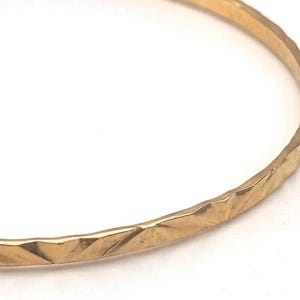 May include: A gold-colored bangle bracelet with a textured, geometric pattern. The bracelet has a simple, elegant design and appears to be made of metal. The surface has a series of diagonal lines, creating a faceted appearance.