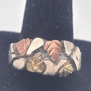 May include: A silver ring with a leaf design. The ring features leaves in gold, rose gold, and silver tones. The leaves are arranged around the band of the ring. The ring is on a black background.