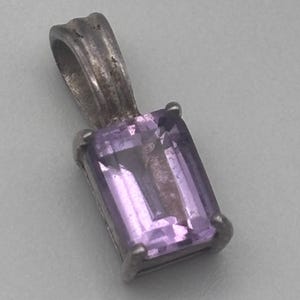 May include: A silver pendant featuring a rectangular, light purple gemstone. The gemstone is set in a silver metal frame with a loop for a chain. The pendant has a simple, elegant design.