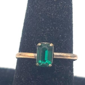 May include: A gold-toned ring featuring a rectangular, emerald-green gemstone. The ring is set on a black display stand, showcasing the vibrant color of the stone. The band appears to be a simple, classic design.