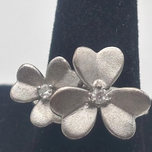 May include: A silver-toned ring featuring two flower-shaped designs. Each flower has five heart-shaped petals and a small, clear gemstone in the center. The ring is displayed against a black background.