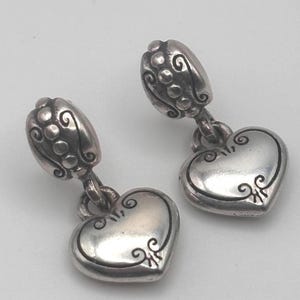 Op de afbeelding: Een paar zilverkleurige oorhangers. Elke oorbel heeft een hartvormige bedel met decoratieve krullen en een bloemvormige bovenkant. De oorbellen hebben een vintage design.